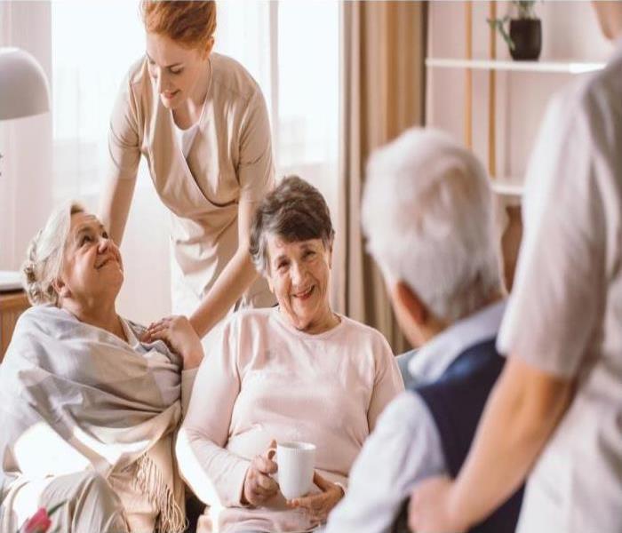 A nurse standing over two elderly women sitting a couch and an elderly man sitting in a wheelchair with a nurse behind him