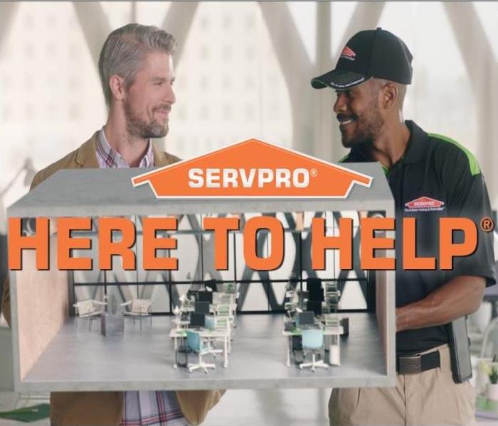 a SERVPRO worker smiling along with business owner, clean office with desk and chairs, with words "Here to Help" in orange. 