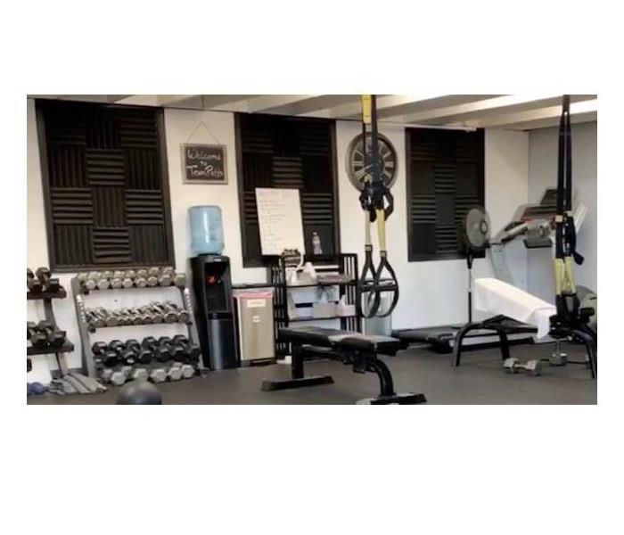 Photo of gym with gray floor with weights against a white wall, water cooler,  and two weight benches.   