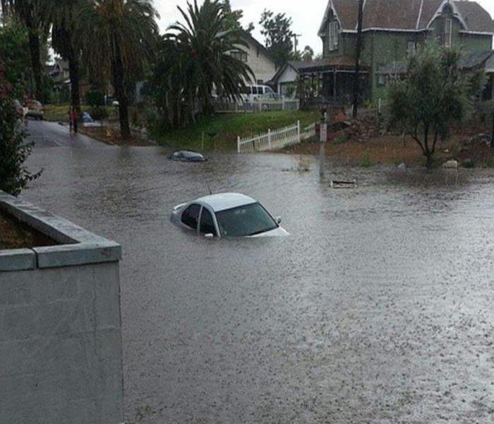 a street flooded by rain water showing a car submerged in the water, with houses in the background